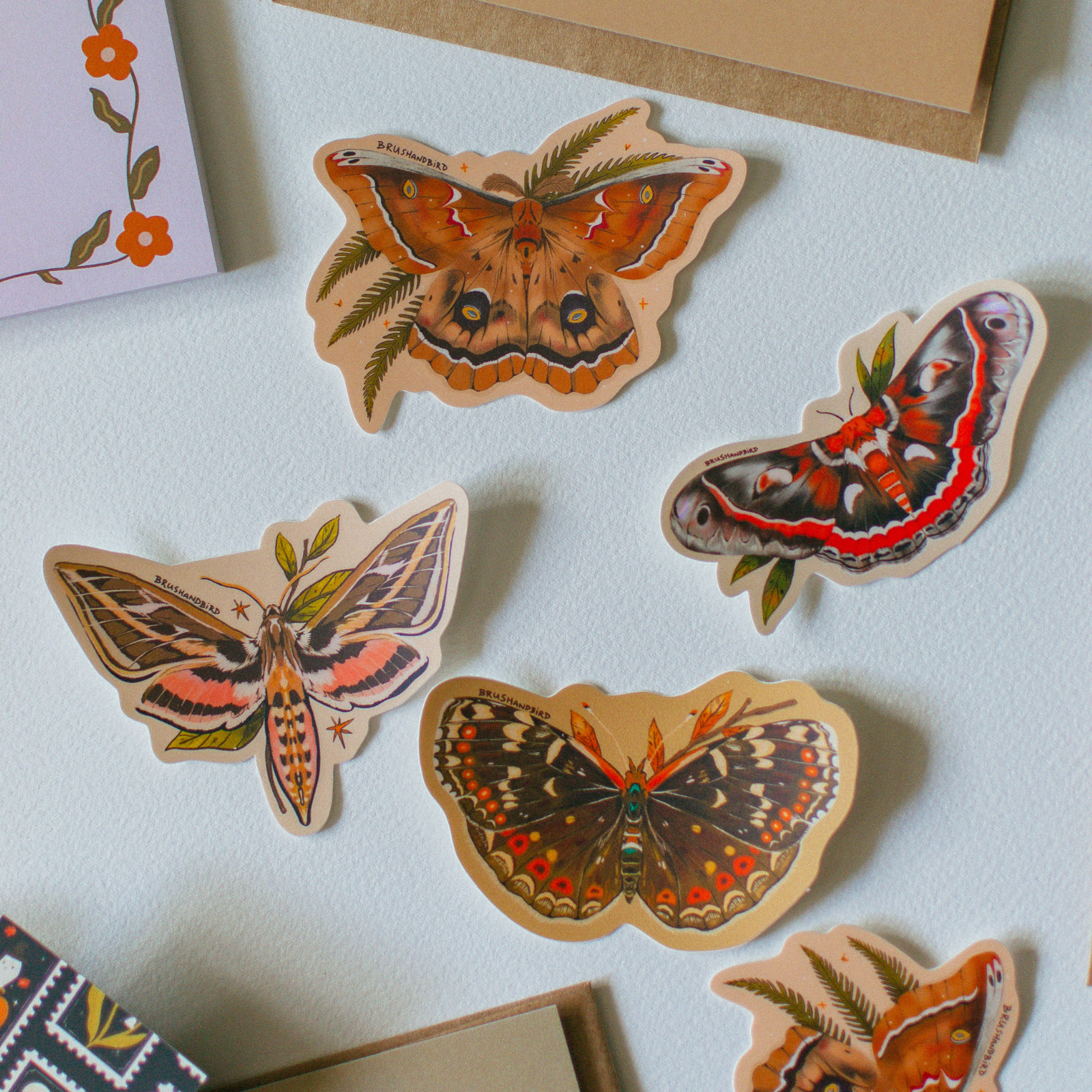 Four die-cut stickers featuring detailed illustrations of various colorful butterflies and moths, arranged on a white textured surface with parts of a kraft envelope and a floral notepad visible.