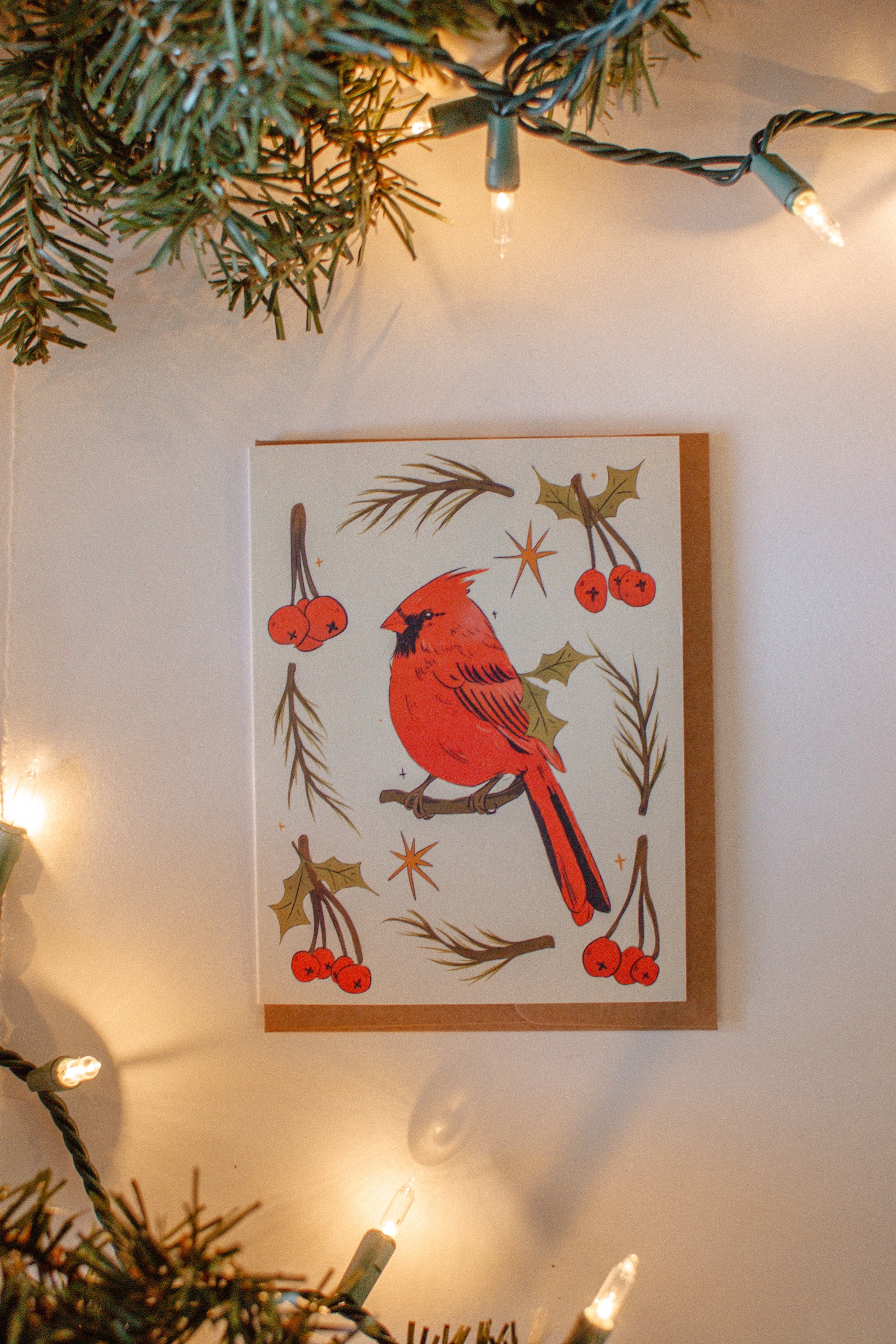 Greeting card of a red cardinal bird with berries on a white background, surrounded by Christmas lights and greenery.