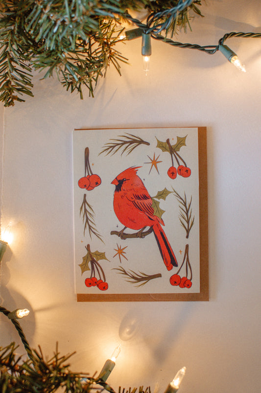 Greeting card of a red cardinal bird with berries on a white background, surrounded by Christmas lights and greenery.