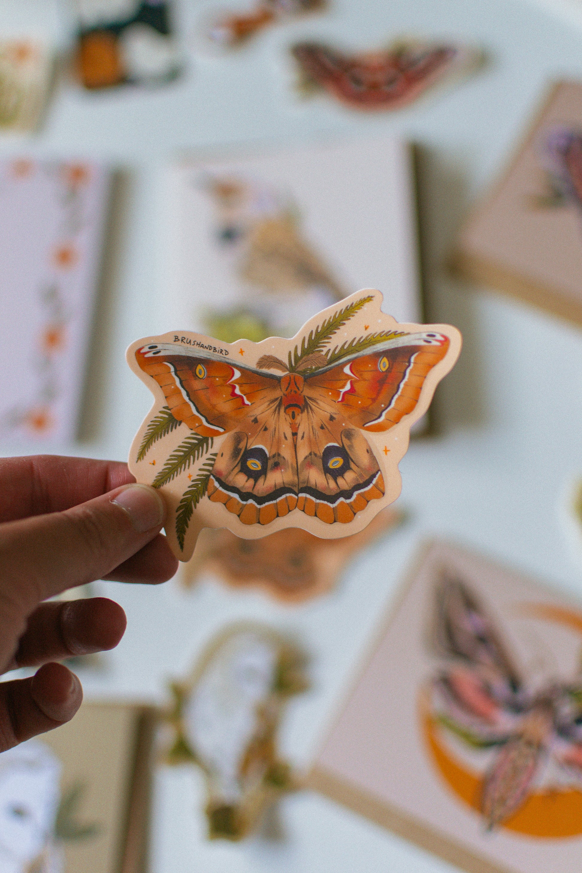 A hand holds a die-cut sticker featuring a detailed illustration of a large moth with orange, brown, and black patterns, set against a softly blurred background of other botanical and insect-themed stickers.
