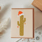 A blank holiday greeting card with a Christmas cactus design, featuring a cactus with a Santa hat and presents, placed on a white surface with holiday decorations around it.