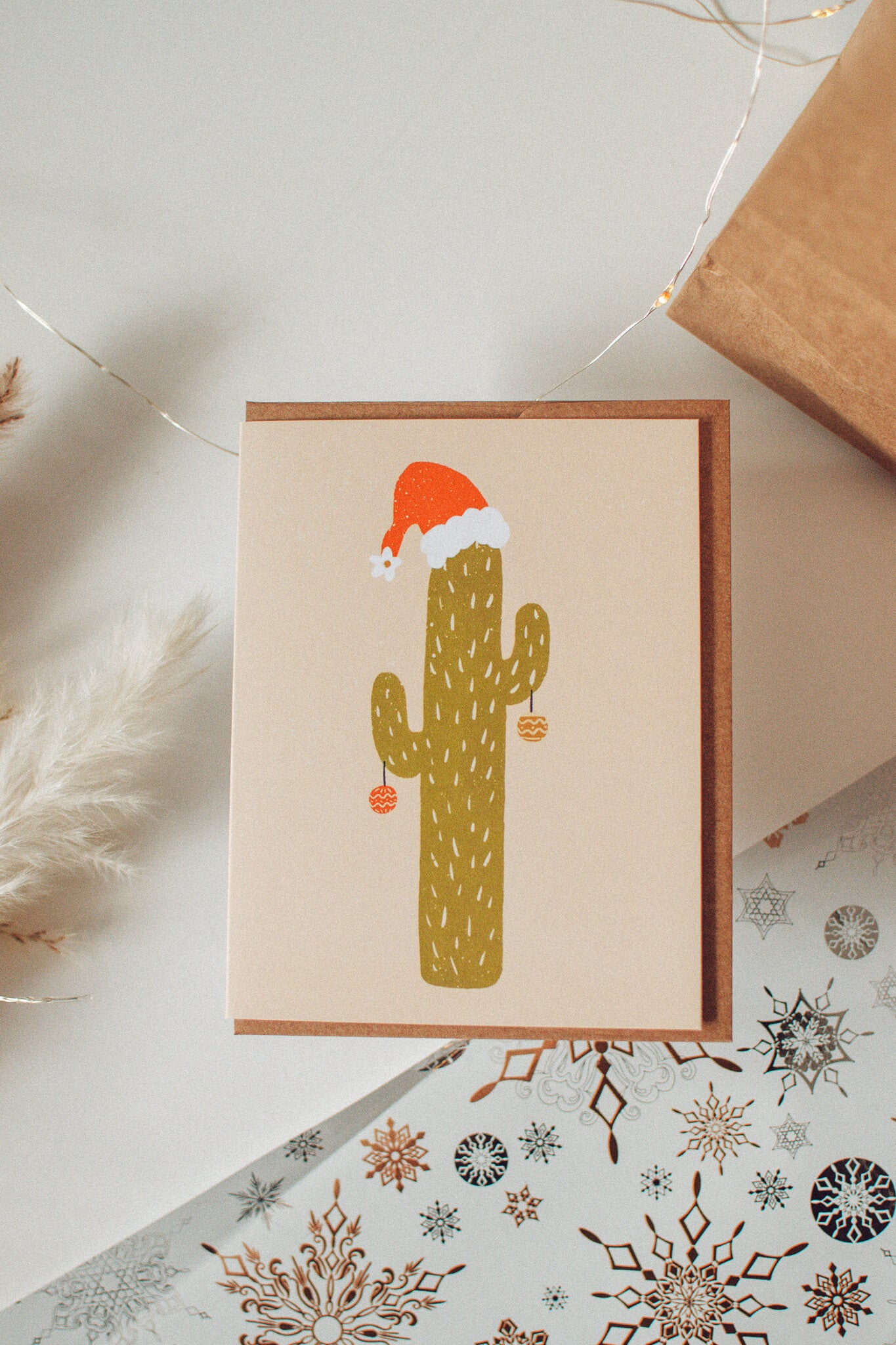 A blank holiday greeting card with a Christmas cactus design, featuring a cactus with a Santa hat and presents, placed on a white surface with holiday decorations around it.