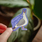 A sticker depicting a seagull with the text 'Menace to Society' printed on it, held between fingers with a plant in the background.