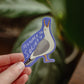 Hand holding a sticker with a seagull design and phrase Menace to Society against a leafy background