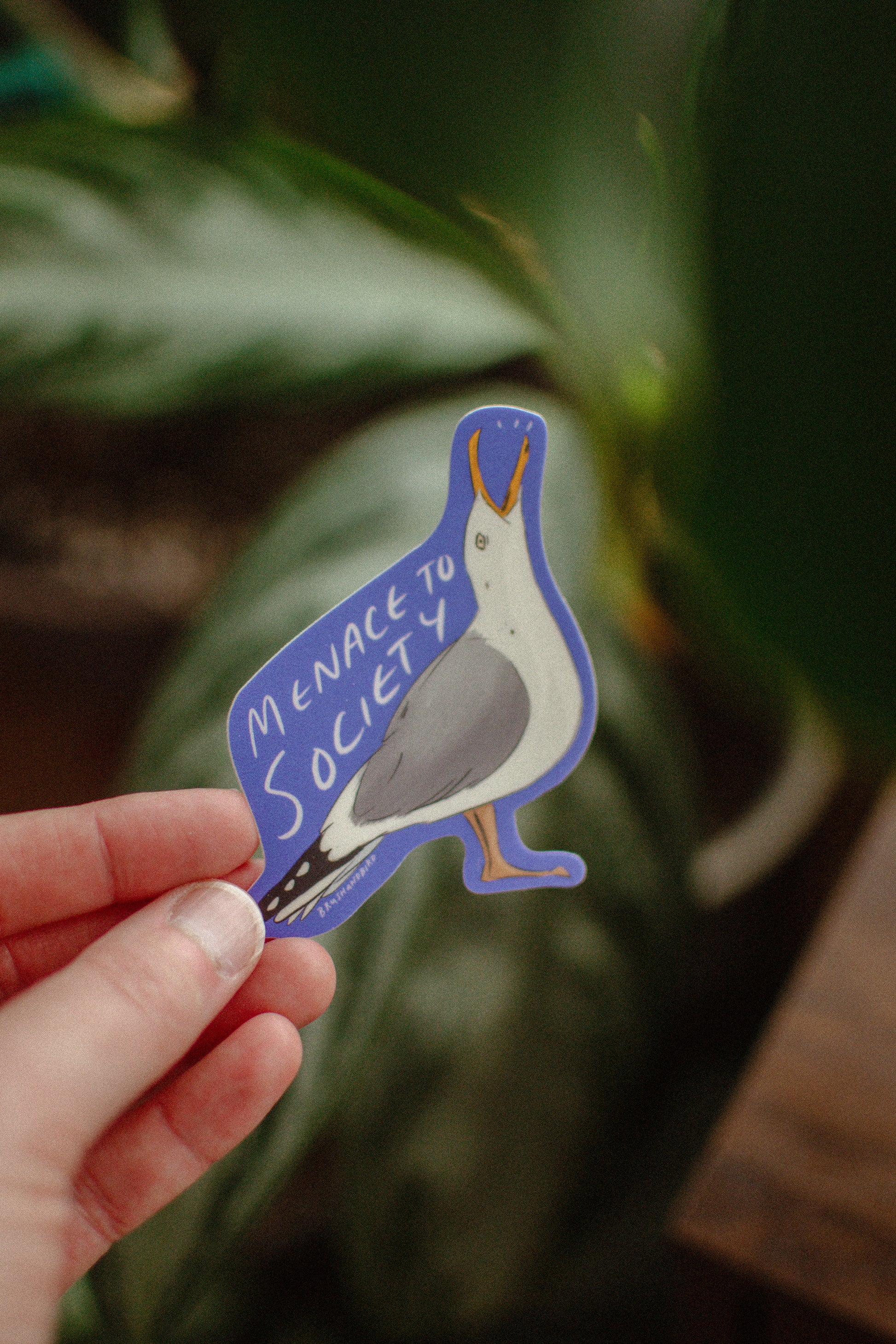 Hand holding a sticker with a seagull design and phrase Menace to Society against a leafy background