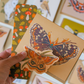 A hand holds a kraft paper card featuring a large illustrated butterfly, with a smaller matching butterfly sticker layered in front. A second card with a mushroom and leaf pattern is visible behind it, and other art cards are blurred in the background.