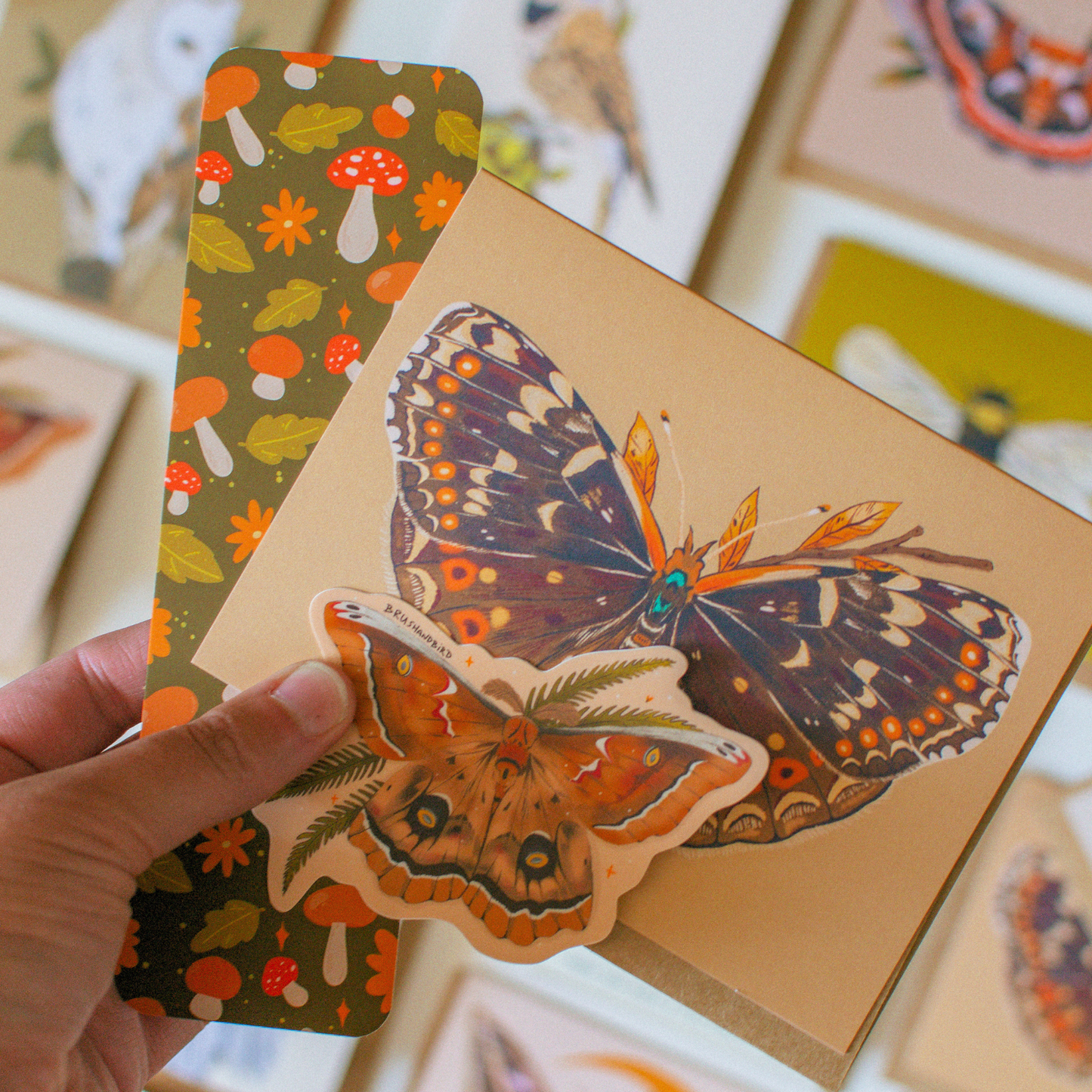 A hand holds a kraft paper card featuring a large illustrated butterfly, with a smaller matching butterfly sticker layered in front. A second card with a mushroom and leaf pattern is visible behind it, and other art cards are blurred in the background.